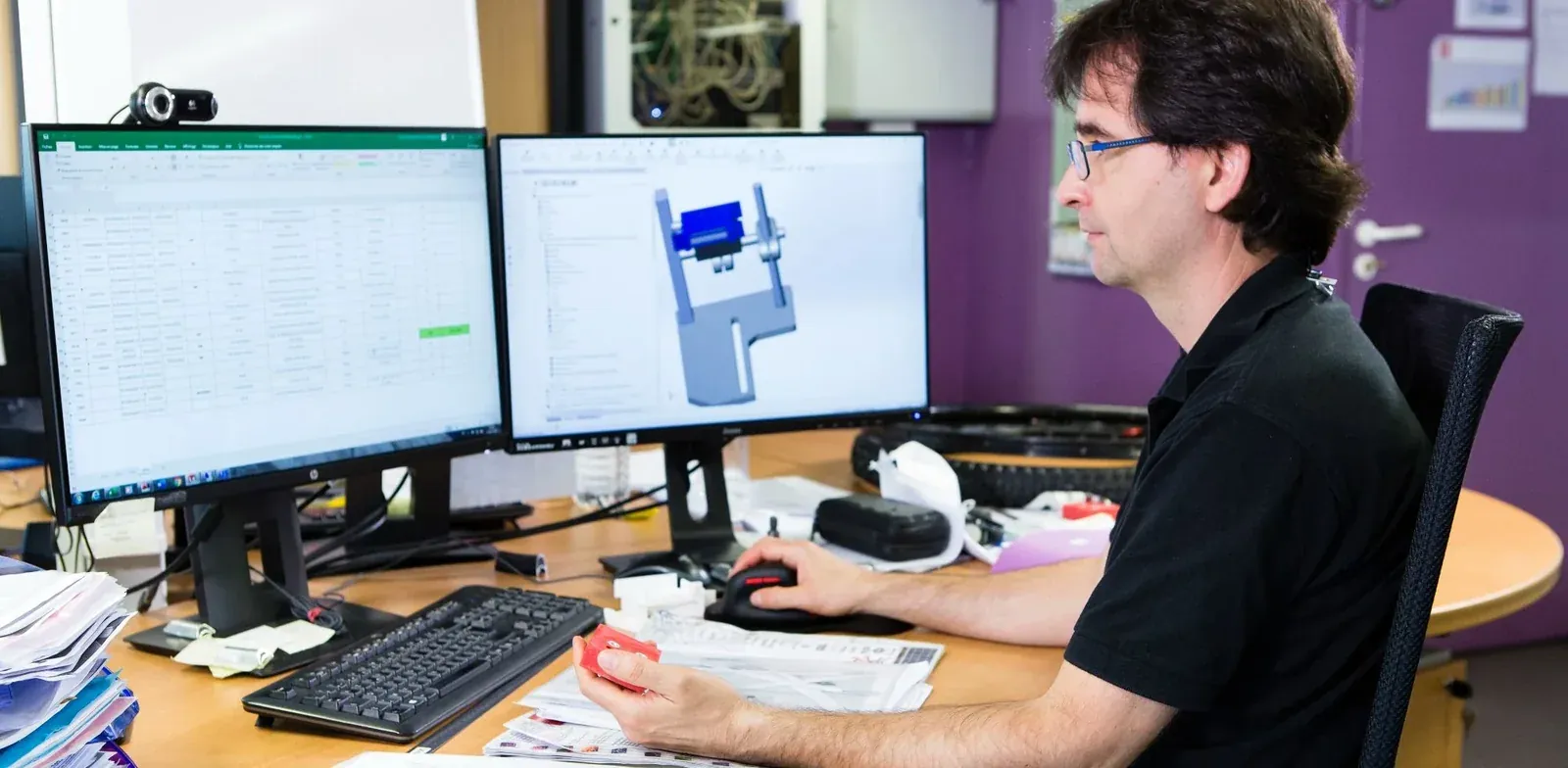 Ingénieur du bureau d'études Mach1 devant deux écrans, un tableur et un plan de conception, une pièce imprimée en 3D à la main