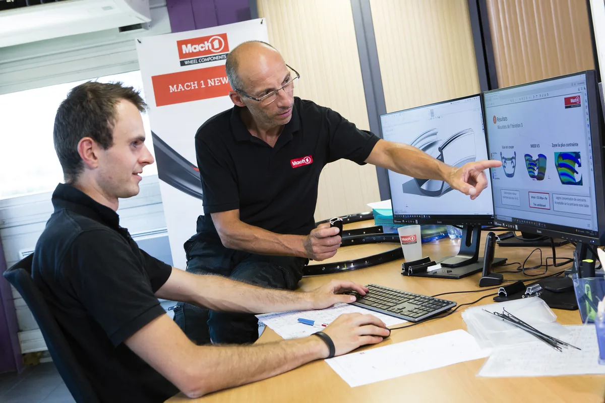 Deux ingénieurs Mach1 devant les écrans du bureau d'études, une jante en main